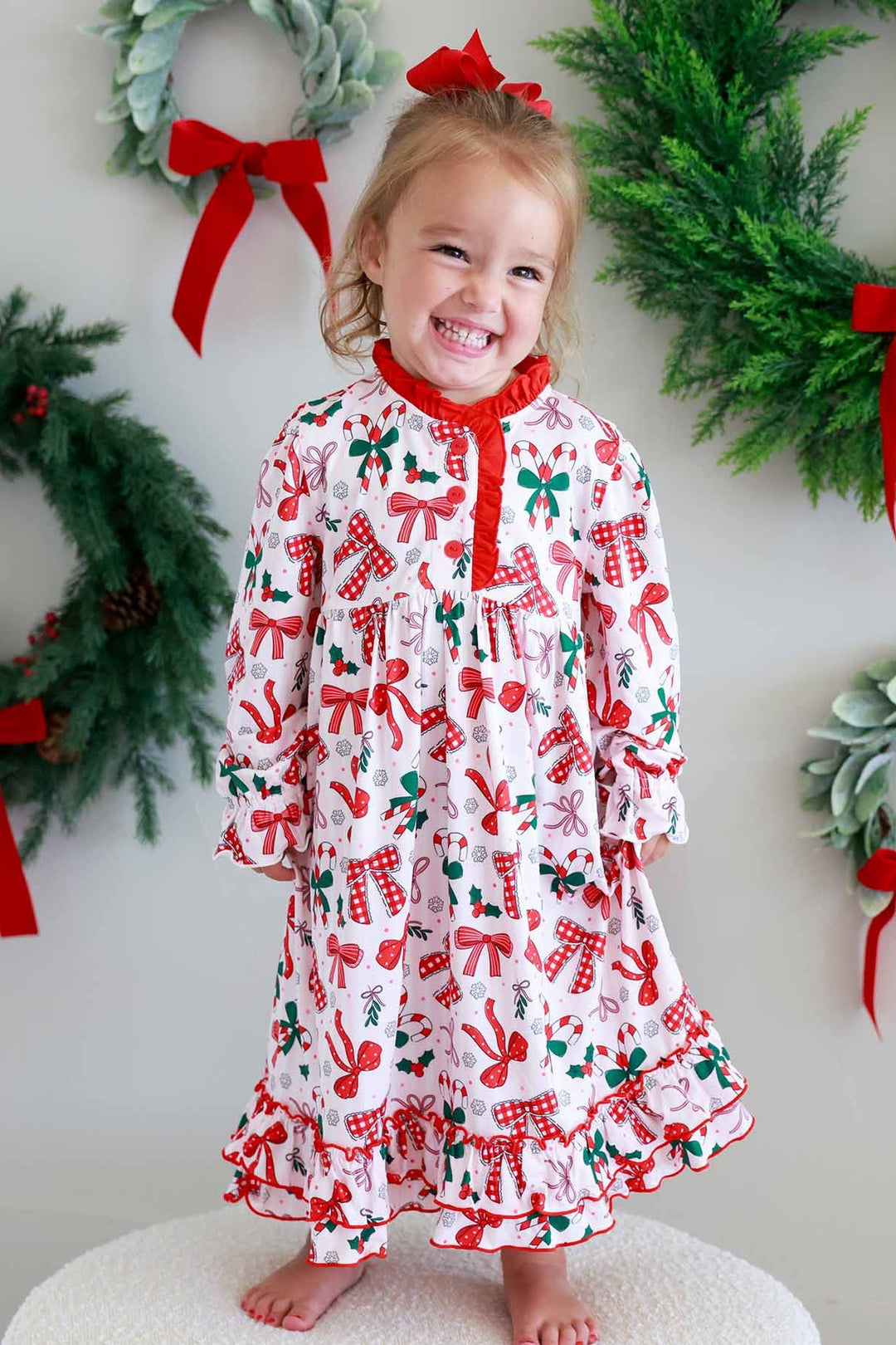 Girl wearing a Caden Lane long-sleeve holiday nightgown with a festive bow, candy cane, and holly print, posing barefoot in front of Christmas wreaths with red bows for a cozy holiday portrait