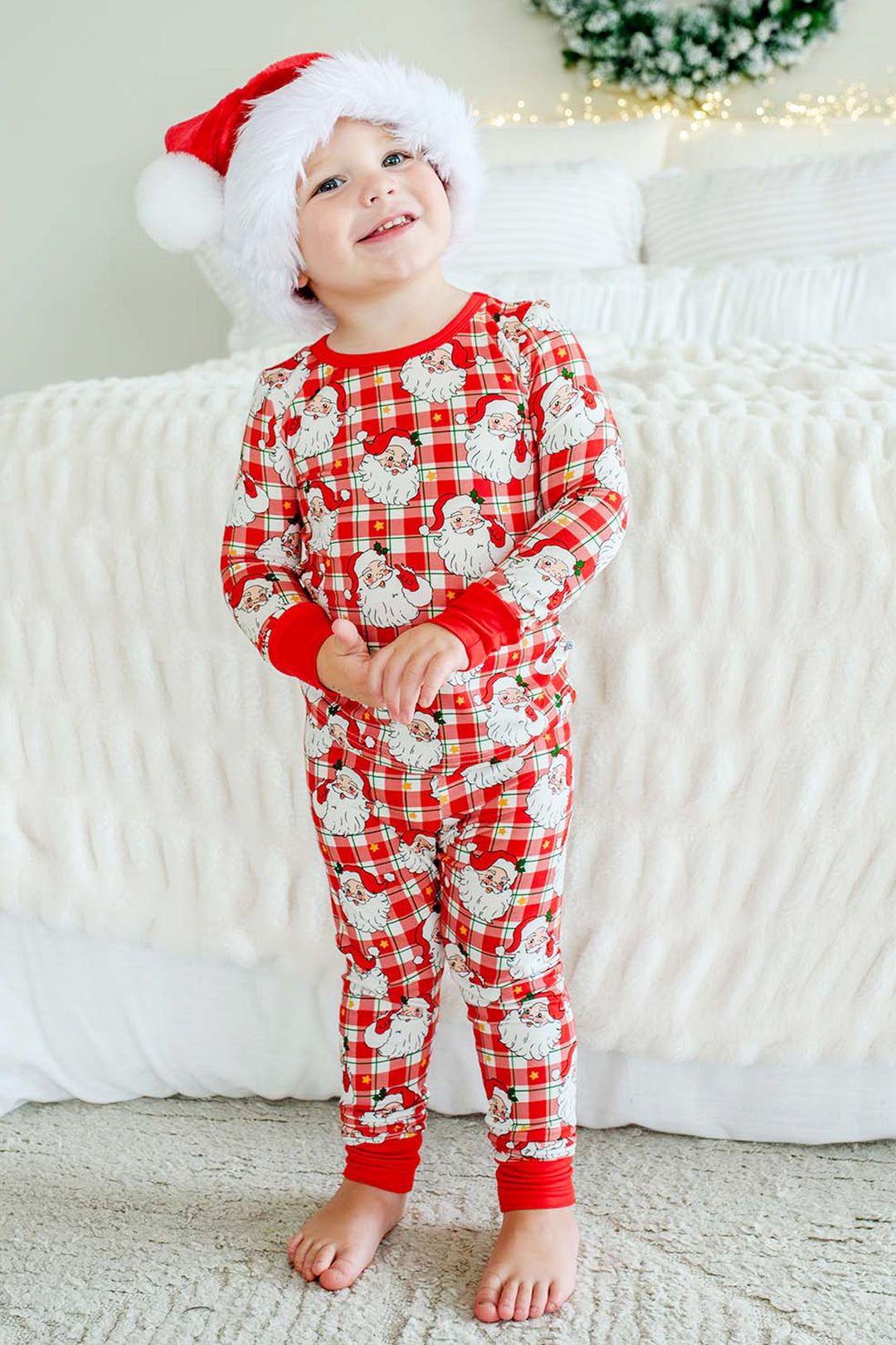 A toddler wearing a red and white Santa plaid pajama set with a white Santa print, standing barefoot in front of a white textured bed and wearing a Santa hat.