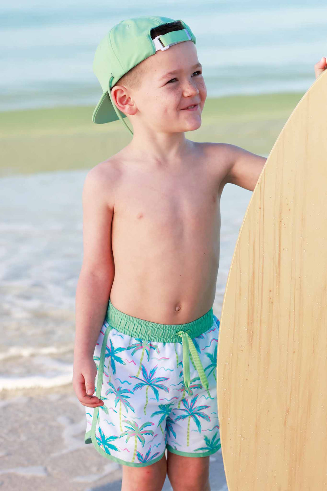 boy standing on beach in palm tree swim trunks with a backwards green surf hat and holding up a wooden surf board