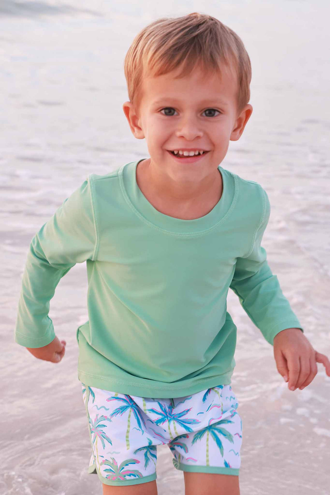 smiling boy in ocean wearing tropical green rash guard shirt with blue lagoon palm tree swim trunks 