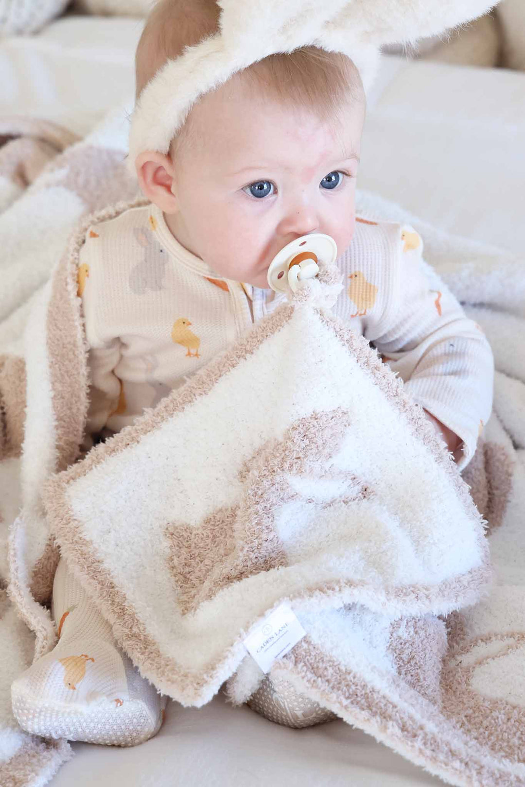 baby with pacifier in mouth with a neutral bunny cuddlelane lovey wearing easter pajamas and bunny ears 