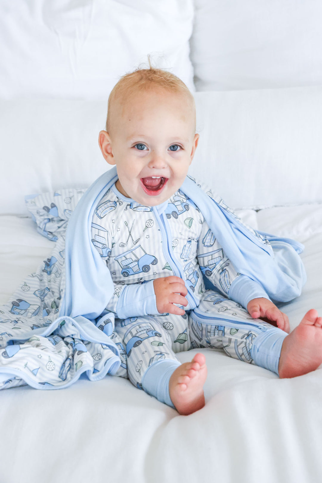 boy sitting on bed in golf themed pajama romper with matching  blanket around shoulders