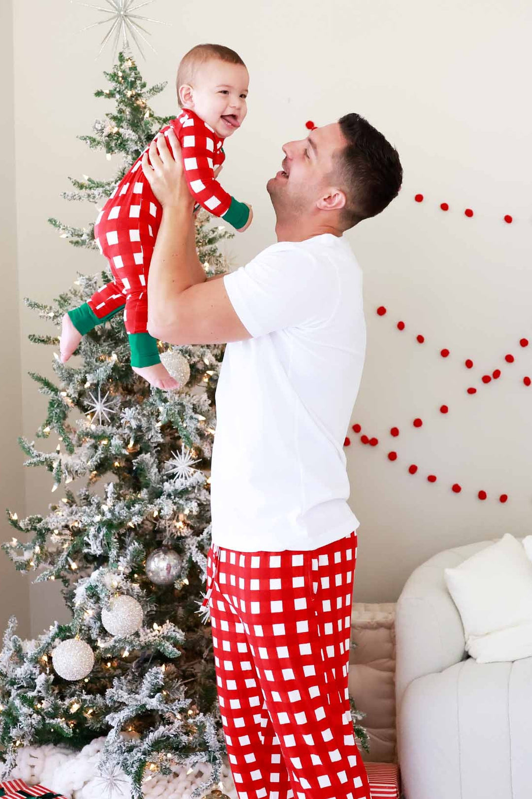 Father and son wearing matching red and white checkered Christmas pajamas, smiling beside a decorated Christmas tree