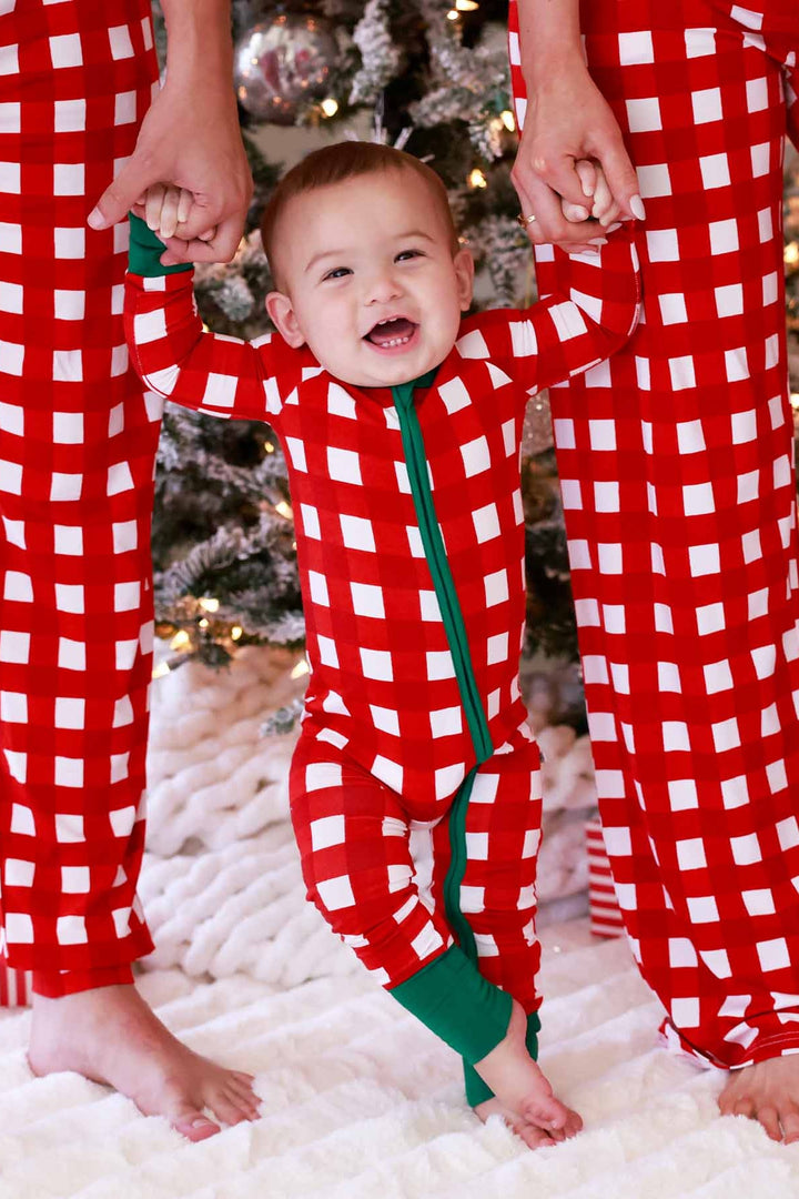 Family of three wearing matching red and white checkered Christmas pajamas, smiling beside a decorated Christmas tree