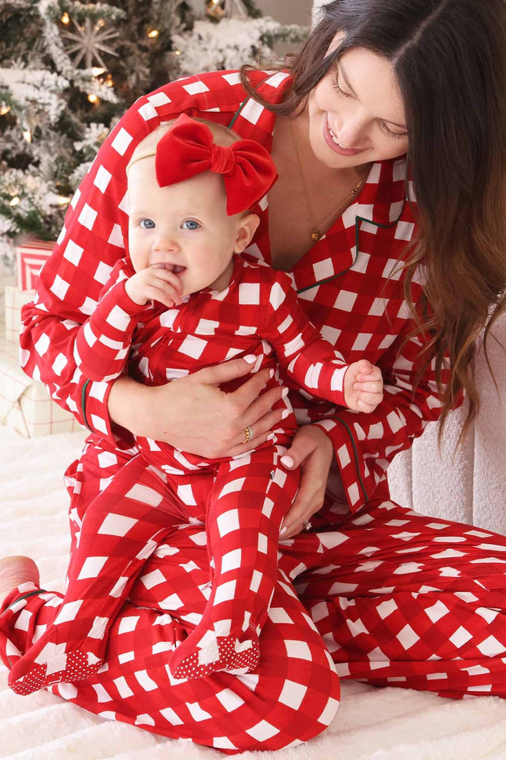 Mother holding her baby while wearing matching Caden Lane red and white checkered holiday pajamas beside a decorated Christmas tree