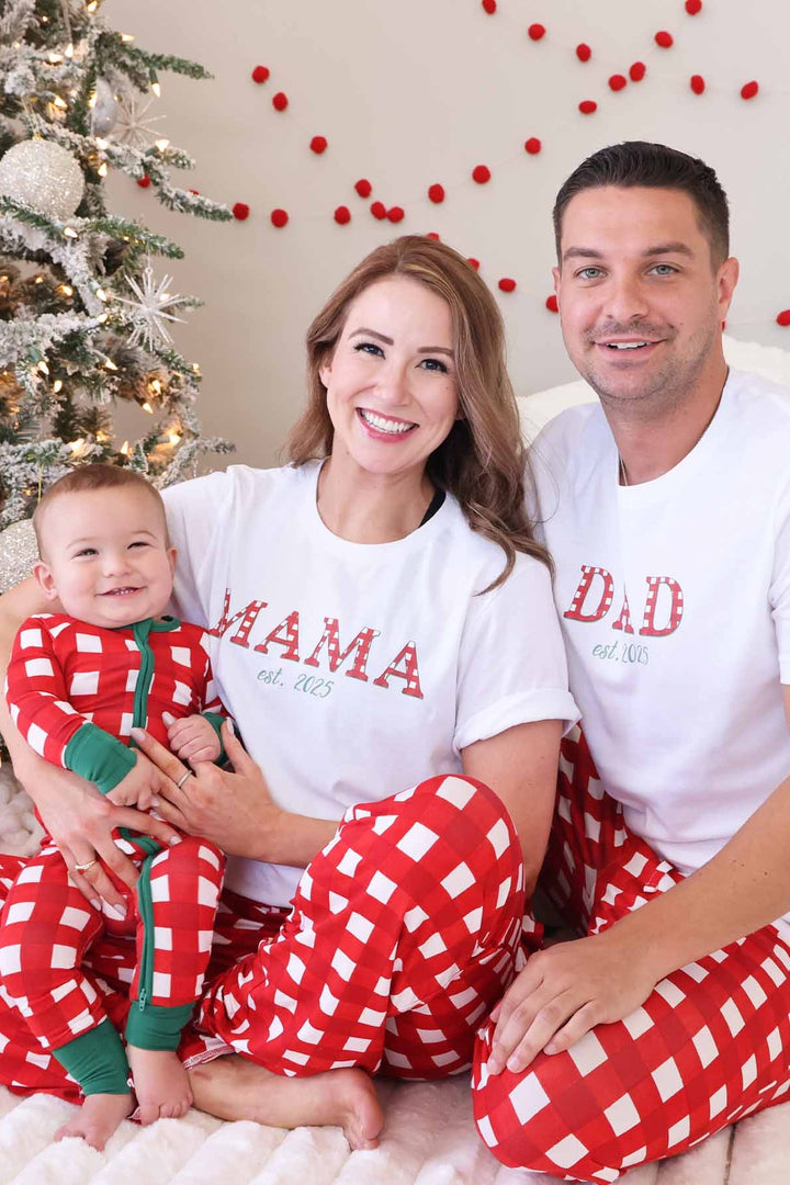 Family of three wearing matching red and white checkered Christmas pajamas, smiling beside a decorated Christmas tree