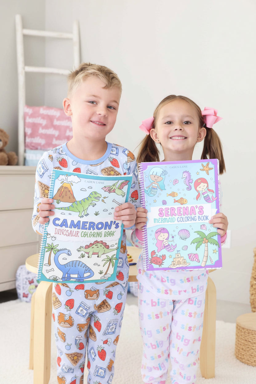 Smiling young boy and girl in pajamas holding their personalized Caden Lane coloring books – 'Cameron's Dinosaur Coloring Book' and 'Serena's Mermaid Coloring Book