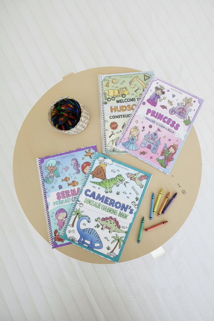 Four personalized Caden Lane coloring books – Hudson's Construction, Aurora's Princess, Serena's Mermaid, and Cameron's Dinosaur – laid out on a wood table with a basket of crayons and loose crayons.