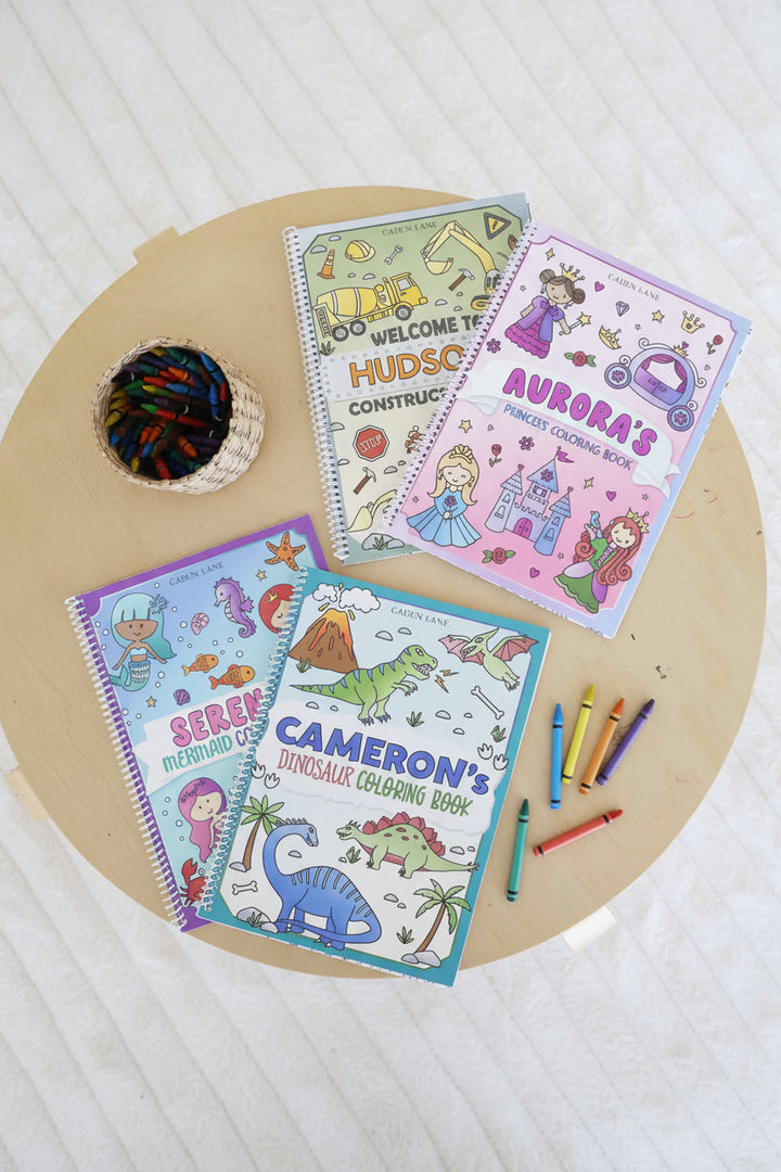 Four personalized Caden Lane coloring books – Hudson's Construction, Aurora's Princess, Serena's Mermaid, and Cameron's Dinosaur – laid out on a wood table with a basket of crayons and loose crayons