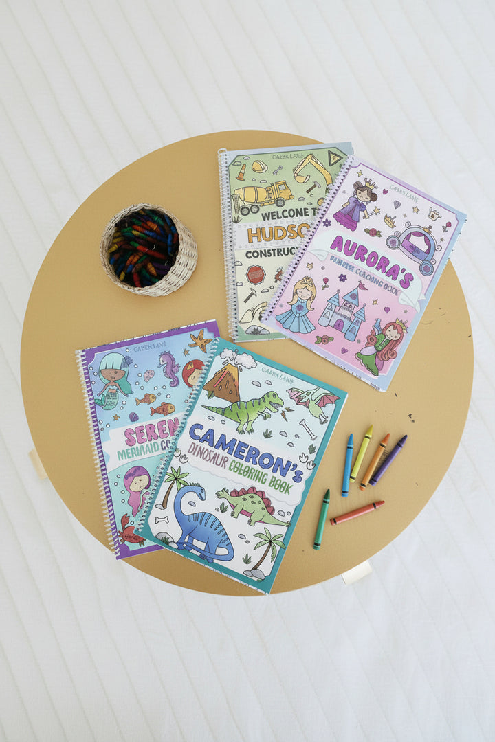 Four personalized Caden Lane coloring books – Hudson's Construction, Aurora's Princess, Serena's Mermaid, and Cameron's Dinosaur – laid out on a wood table with a basket of crayons and loose crayons.