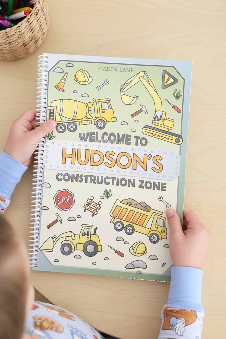 Child's hands holding open Caden Lane 'Welcome to Hudson's Construction Zone' coloring book, showing cement truck and excavator illustrations