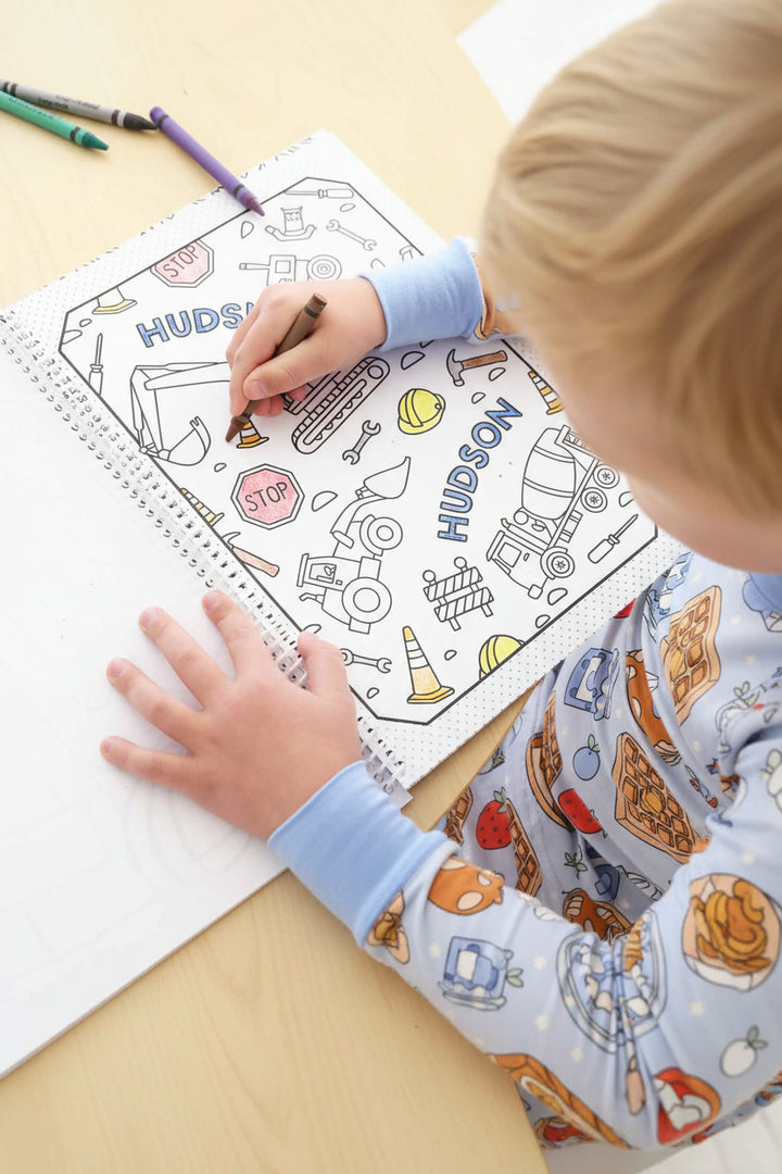 Child coloring a personalized 'Hudson' construction-themed name page with a brown crayon, filling in an excavator illustration in a Caden Lane coloring book