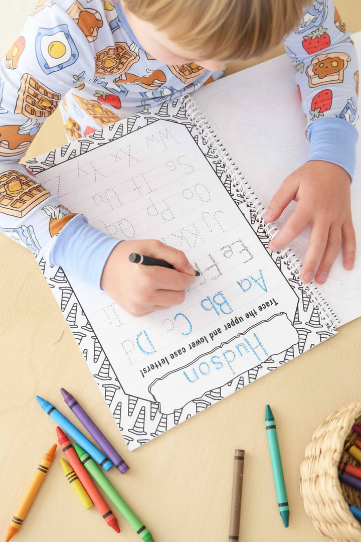 Child tracing personalized 'Hudson' alphabet letters with a black crayon on a construction cone-bordered practice page in a Caden Lane coloring book