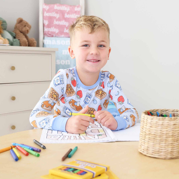 Smiling young boy in a waffle-print pajama set coloring a construction-themed page with a yellow crayon at a wooden table, surrounded by crayons and a crayon basket