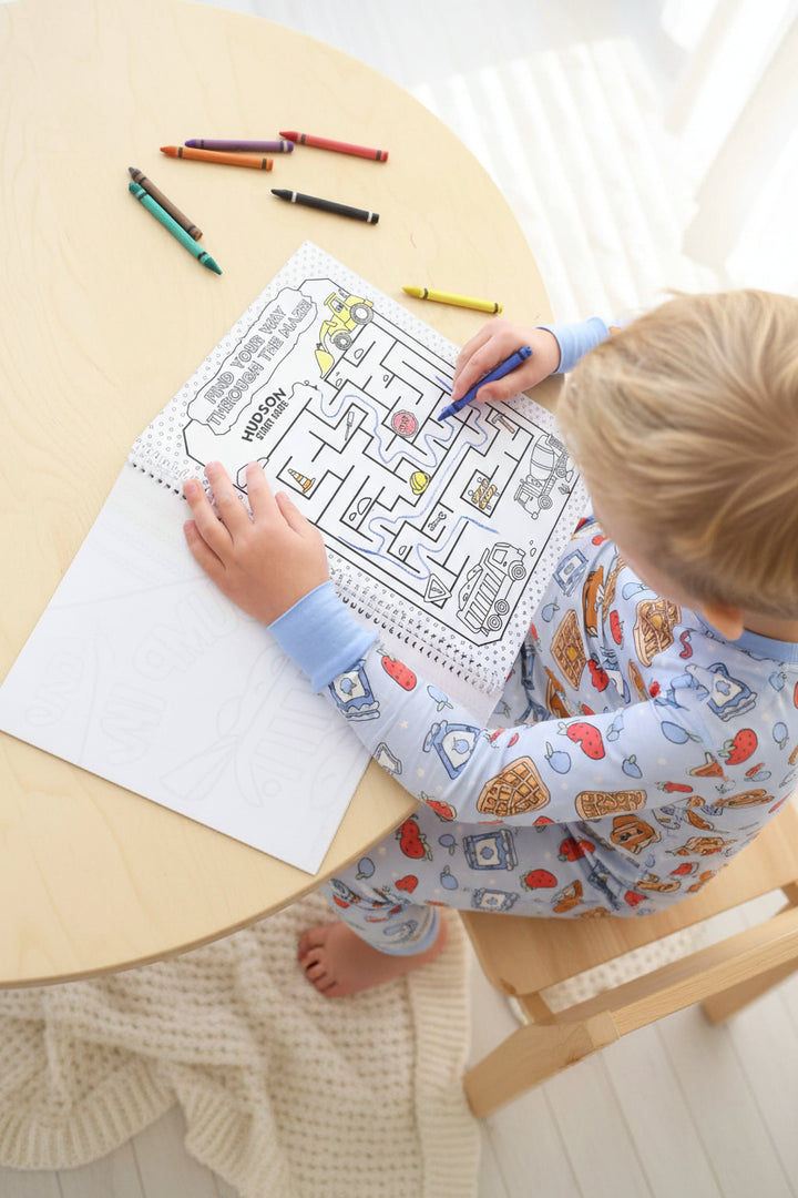 Child coloring a personalized 'Hudson, start here' maze page with a blue crayon, navigating construction vehicle illustrations in a Caden Lane coloring book.
