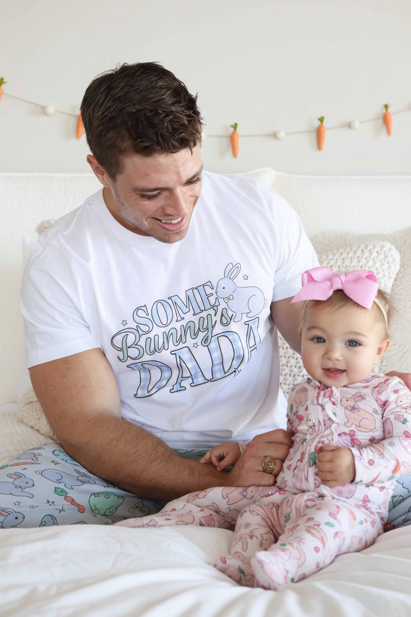 dad wearing the some bunny's dada easter short sleeve graphic tee with patching pajama pants holding a baby girl wearing a pink ruffle zipper footie 