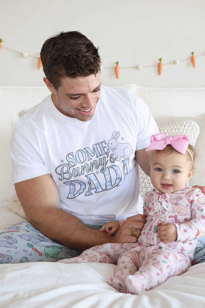 dad wearing the some bunny's dada easter short sleeve graphic tee with patching pajama pants holding a baby girl wearing a pink ruffle zipper footie 