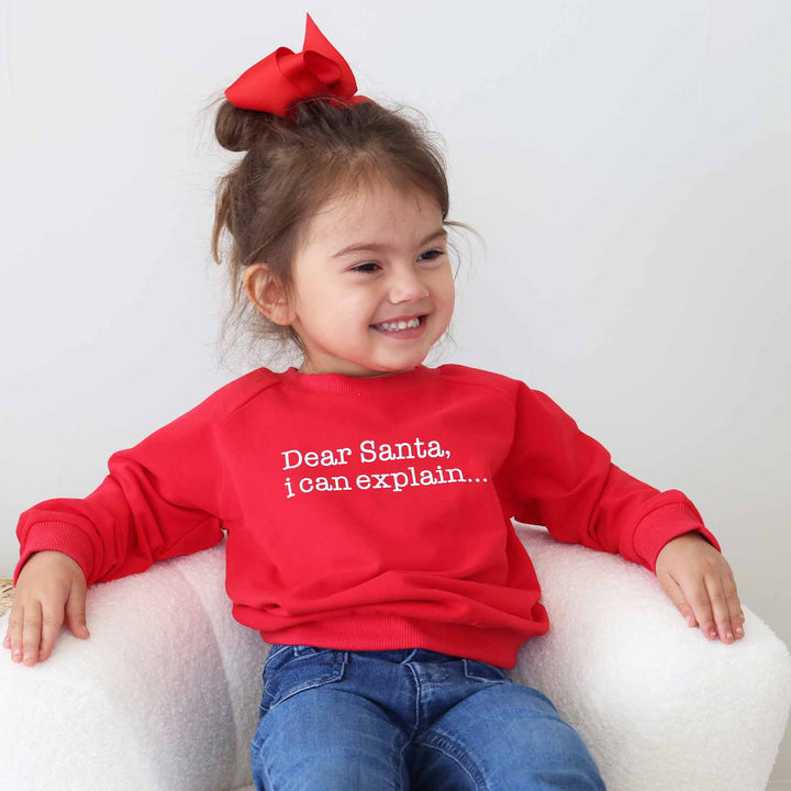 kid sitting back in chair wearing a long sleeve red sweatshirt that says "dear santa i can explain"