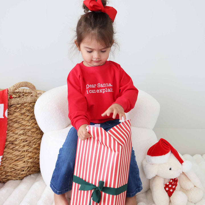 little girl opening presents wearing jeans and a long sleeve red sweatshirt that says "dear santa i can explain"