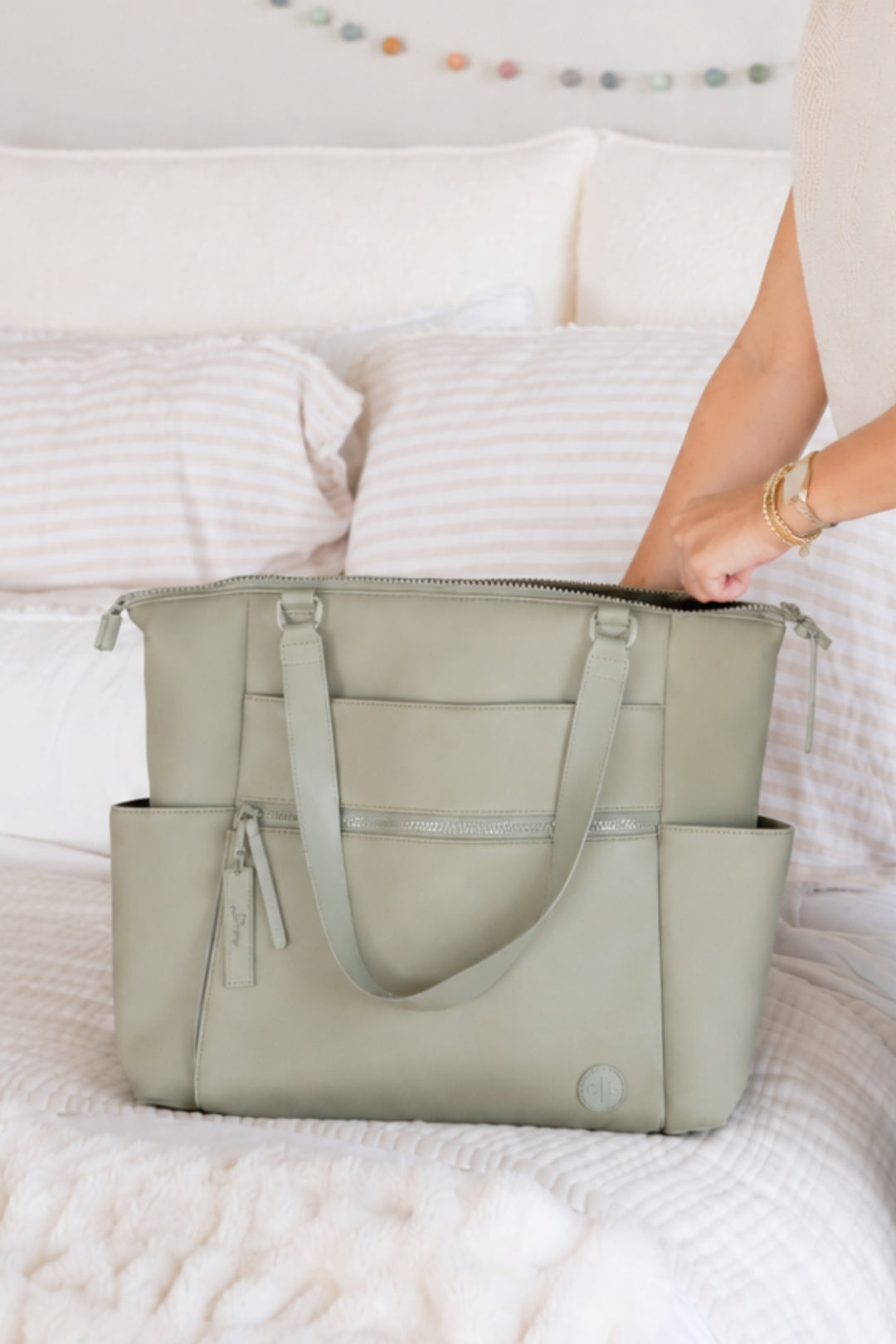 A sage green diaper bag with dual shoulder straps and zippered compartments, shown on a white bed with a person's arm reaching in from the right.