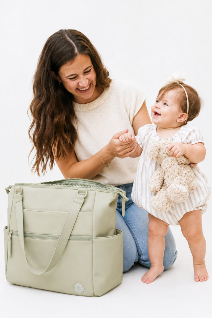 A woman kneeling on a white studio floor smiles while holding a baby, with a large sage green diaper bag beside her. The baby wears a white and beige striped romper and holds a beige teddy bear.