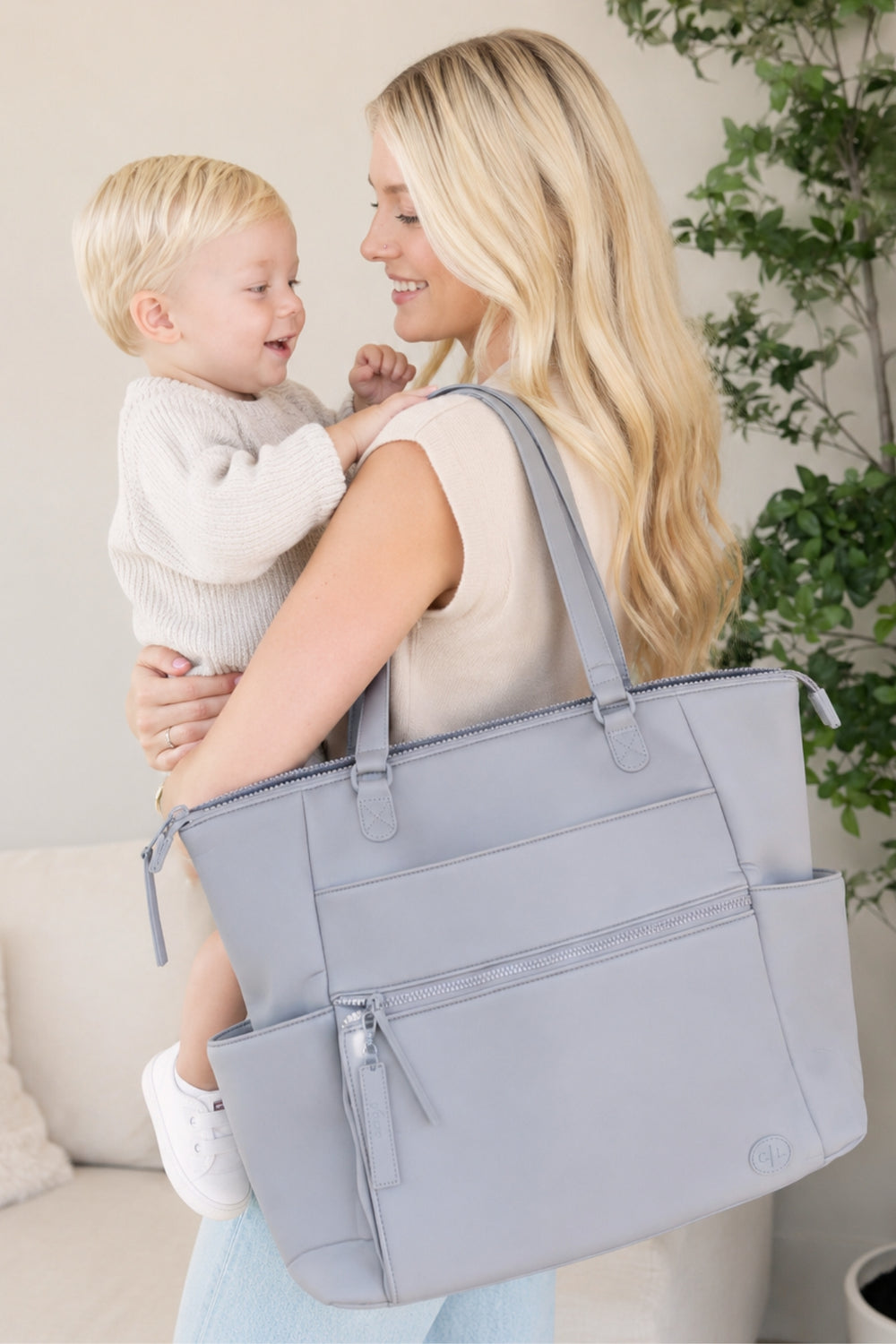 A woman carrying a young child over her shoulder in a bright indoor setting, holding a large light blue-gray diaper bag with multiple zip compartments and side pockets.