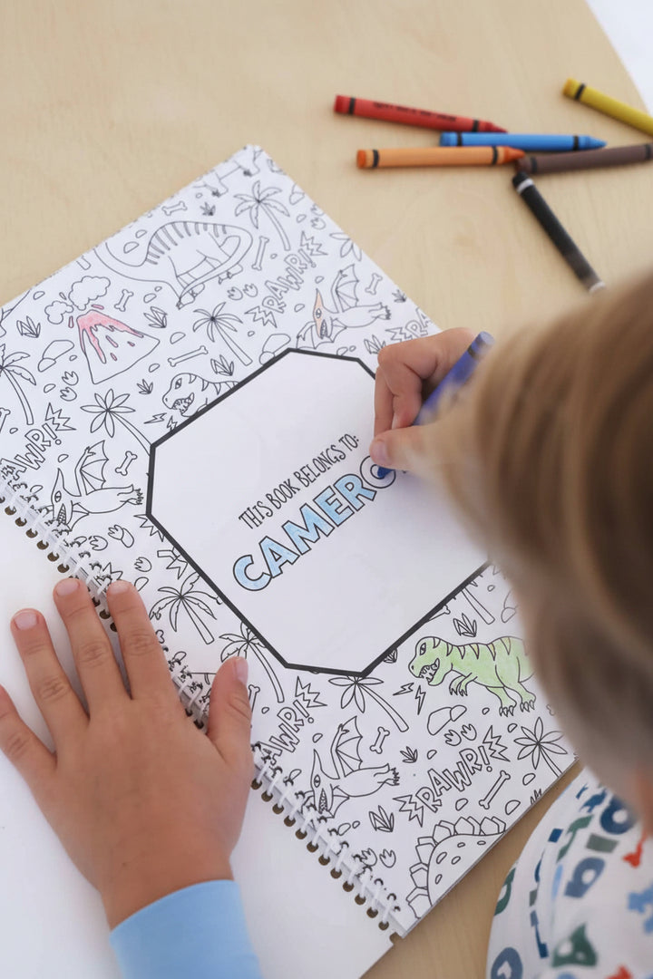 Child in a Big Brother pajama set coloring the 'This Book Belongs To: Cameron' title page with a blue crayon, surrounded by dinosaur and volcano illustrations in a Caden Lane coloring book
