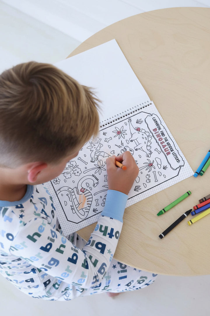 Child in a Big Brother pajama set coloring a personalized 'Cameron, find the hidden letters: DINOSAUR' activity page with an orange crayon in a Caden Lane coloring book