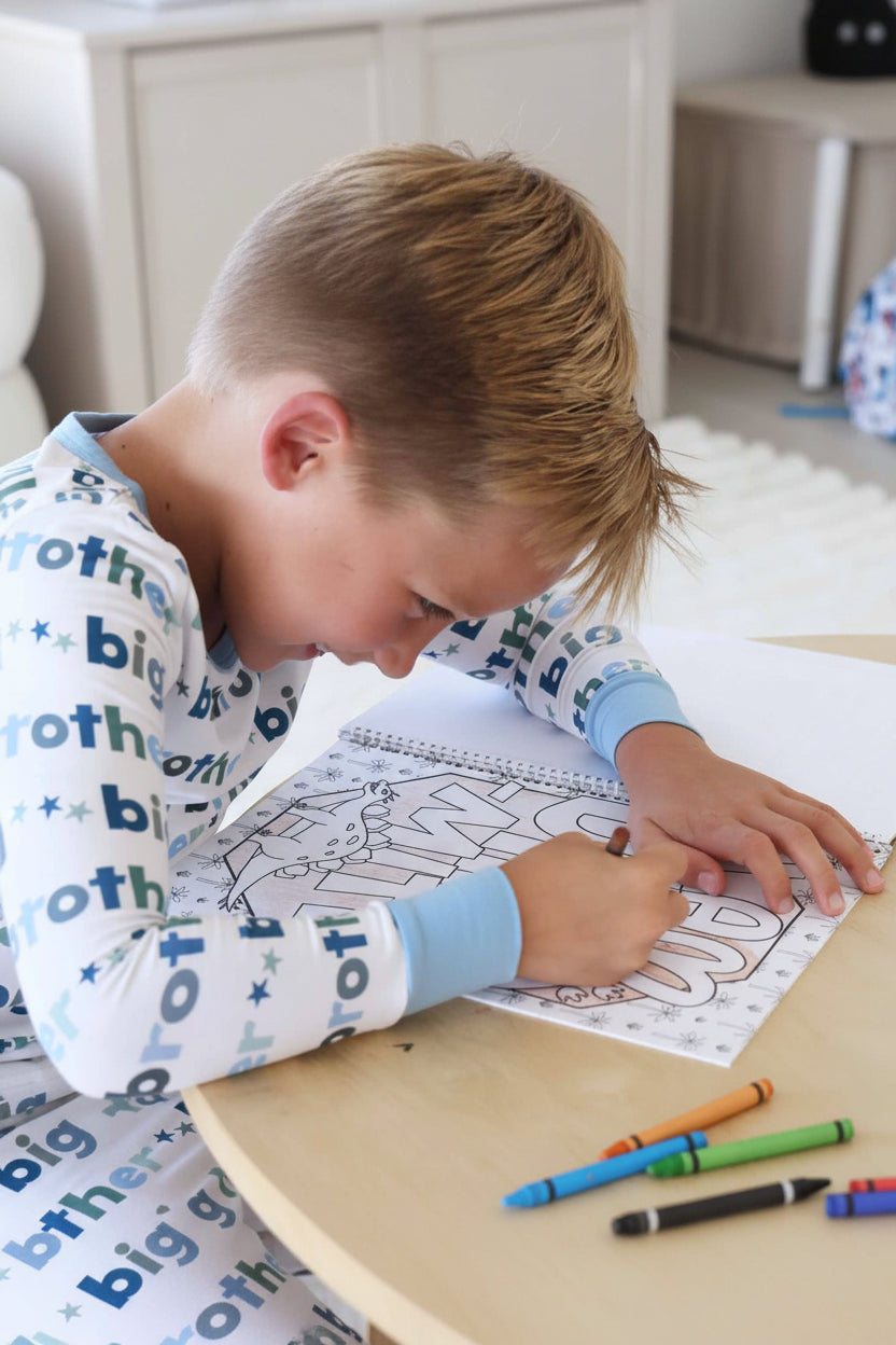 Child in a Big Brother pajama set coloring a 'My name is' page with a brown crayon, filling in a dinosaur illustration in a Caden Lane coloring book
