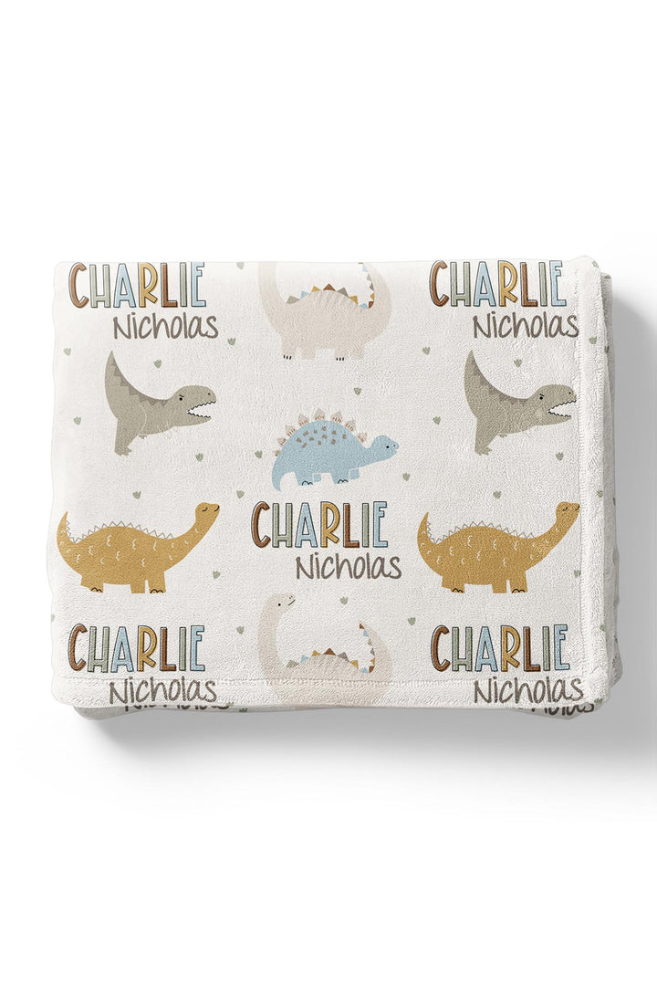 Flat lay of a folded personalized Caden Lane Dino Stomps blanket in cream, showing dinosaur silhouettes, hearts, and custom name print