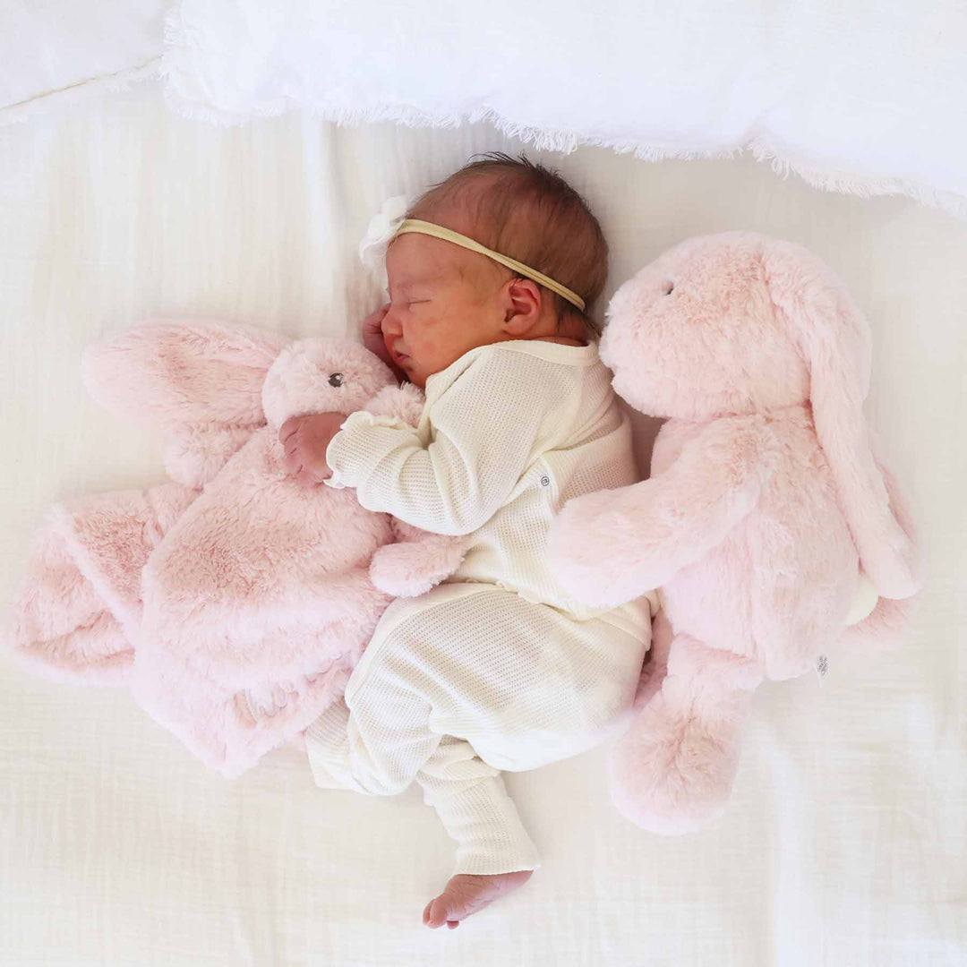 baby laying on side with pink bunny stuffed animal behind her holding a pink embroidered bunny lovey