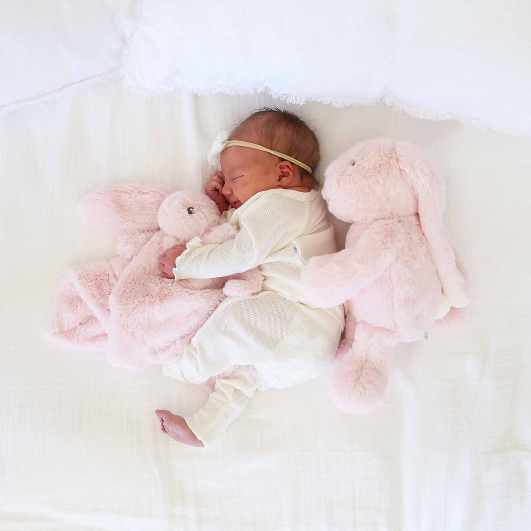 baby laying on side holding a light pink stuffed animal lovey for babies with a pink bunny stuffed animal behind her 