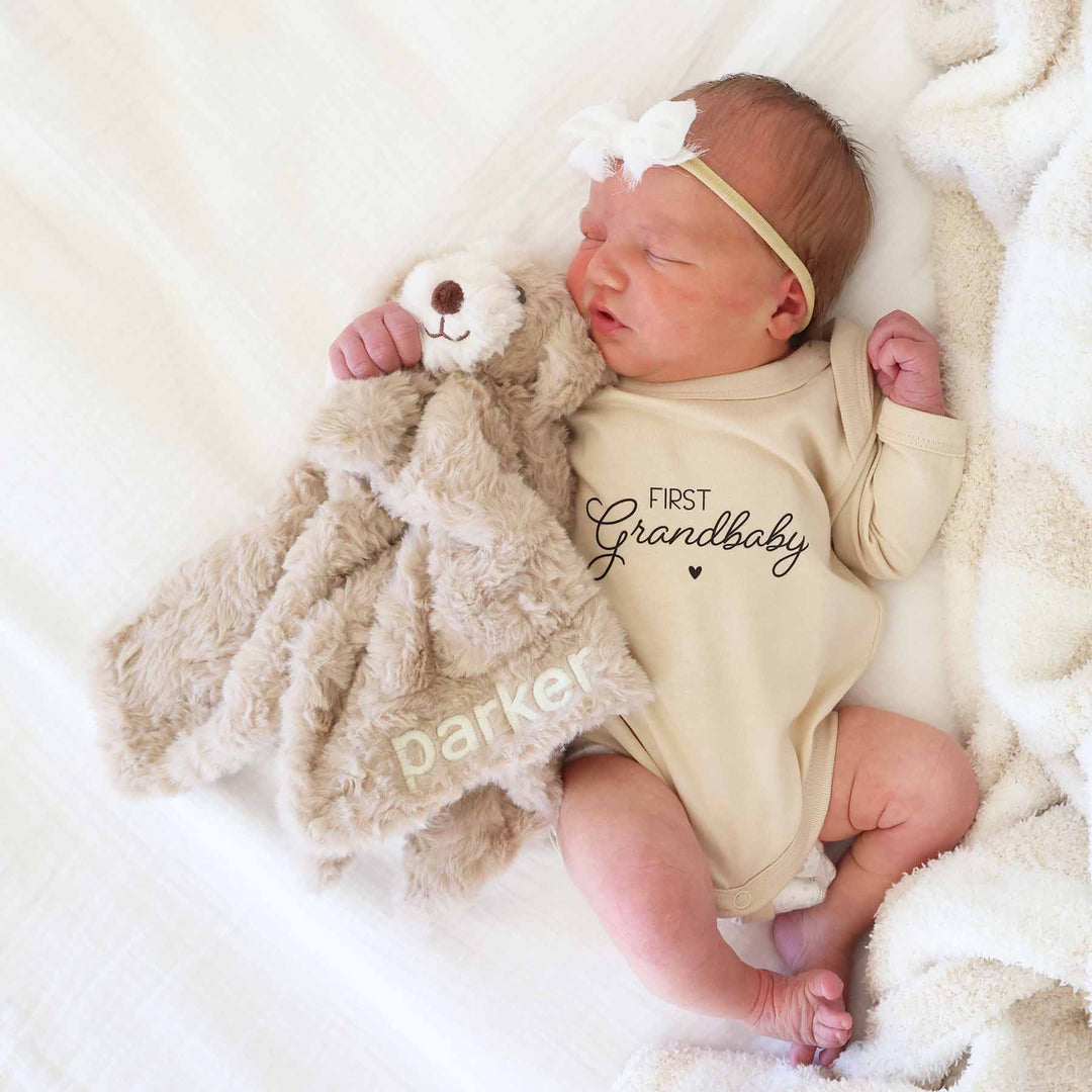 baby laying on bed in first grandbaby graphic bodysuit holding an embroidered puppy lovey