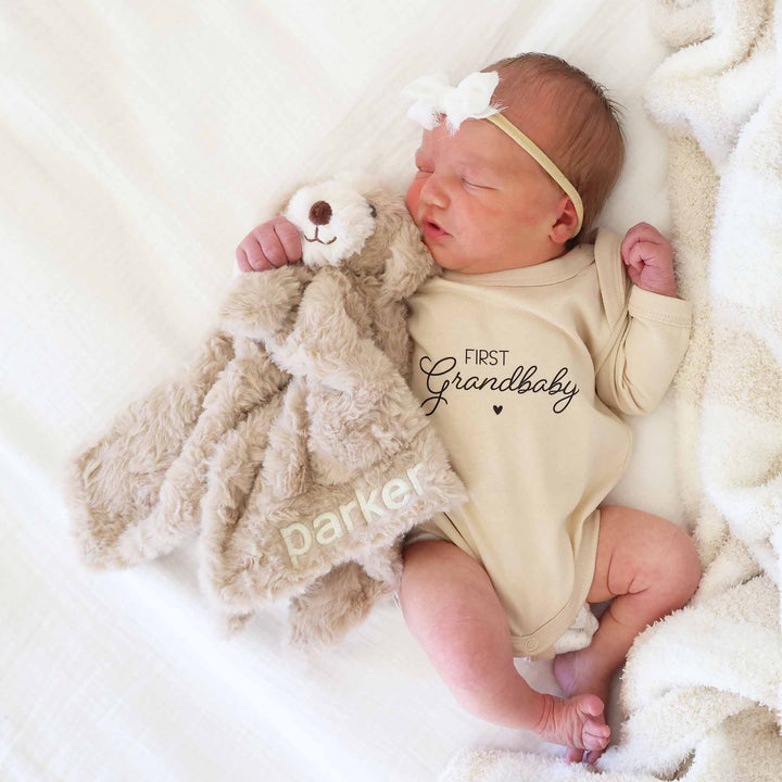 baby laying on bed in first grandbaby graphic bodysuit holding an embroidered puppy lovey