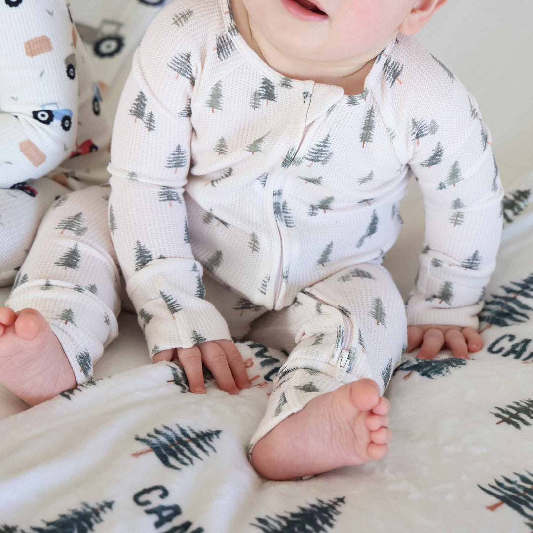 baby sitting and wearing the evergreen controvertible zip romper. Green pine trees with white background 