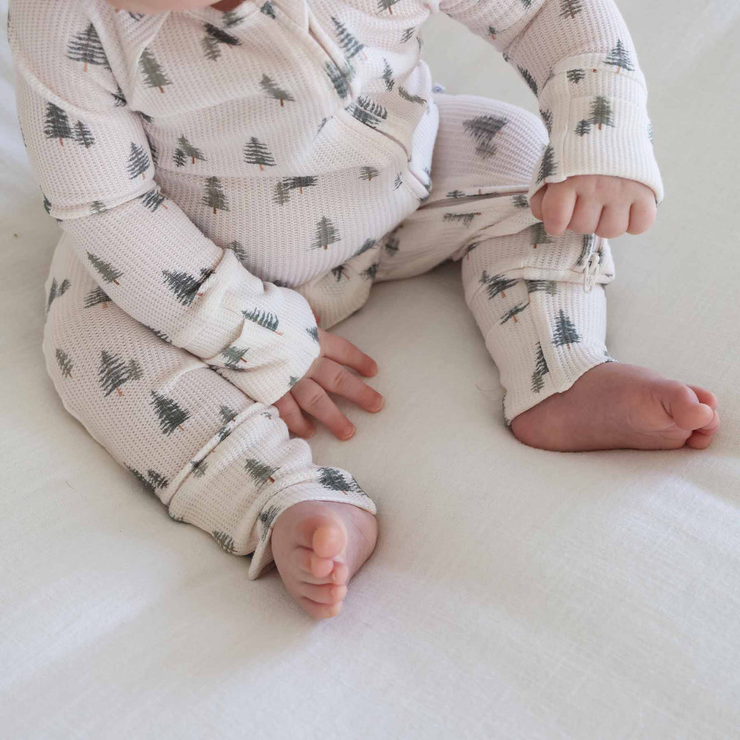 close up of baby sitting and wearing the evergreen controvertible zip romper. Green pine trees with white background 