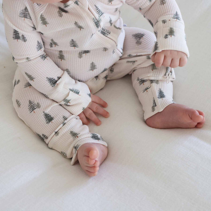 close up of baby sitting and wearing the evergreen controvertible zip romper. Green pine trees with white background 