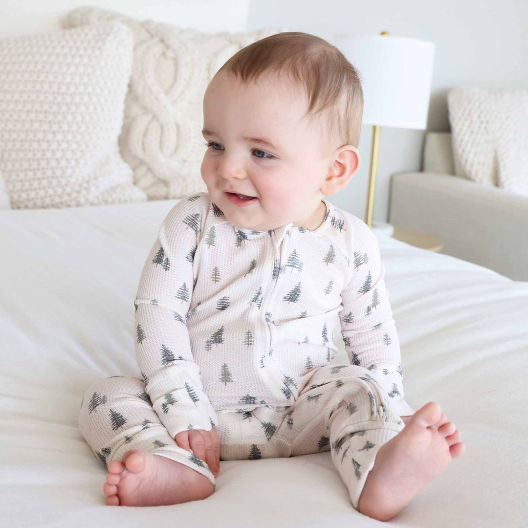 baby sitting and wearing the evergreen controvertible zip romper. Green pine trees with white background 