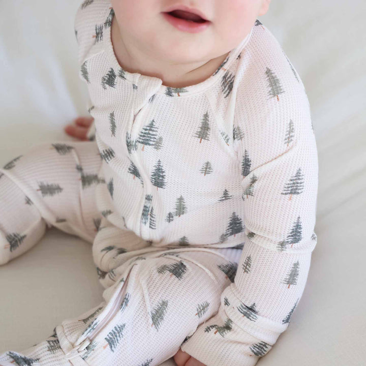 close up of baby sitting and wearing the evergreen controvertible zip romper. Green pine trees with white background 
