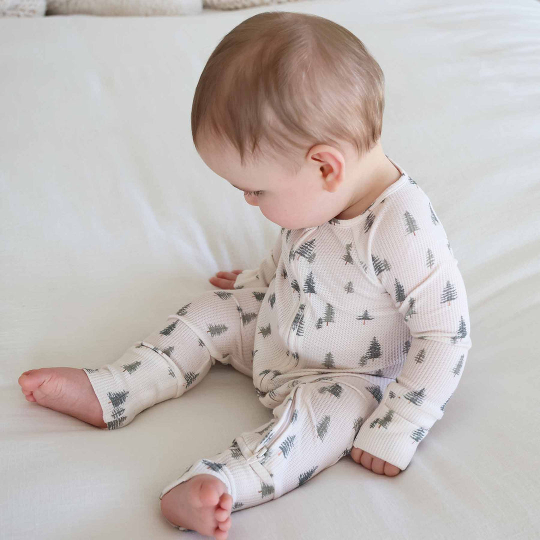 baby sitting and wearing the evergreen controvertible zip romper. Green pine trees with white background 