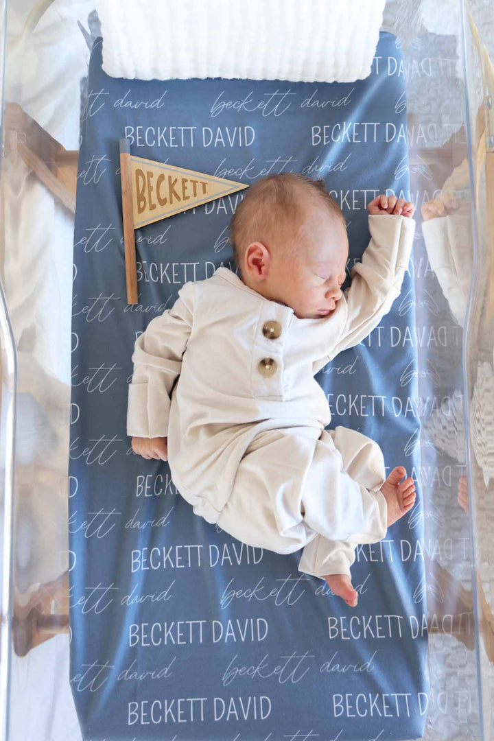 newborn boy laying on blue personalized swaddle blanket in a white two piece outfit with a flag personalized wooden sign next to him
