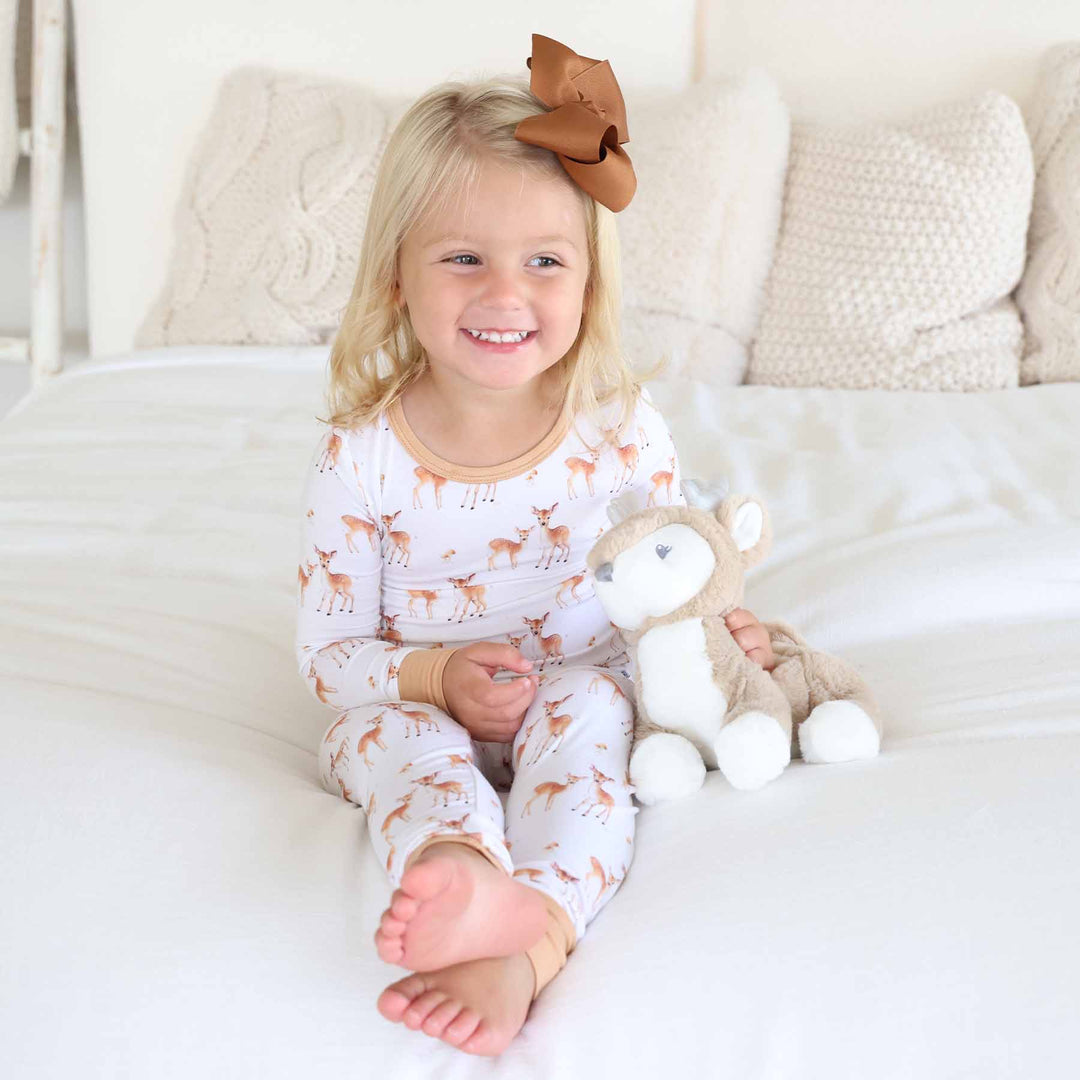 Smiling toddler in fawn print pajamas holding a plush deer on a cozy bed