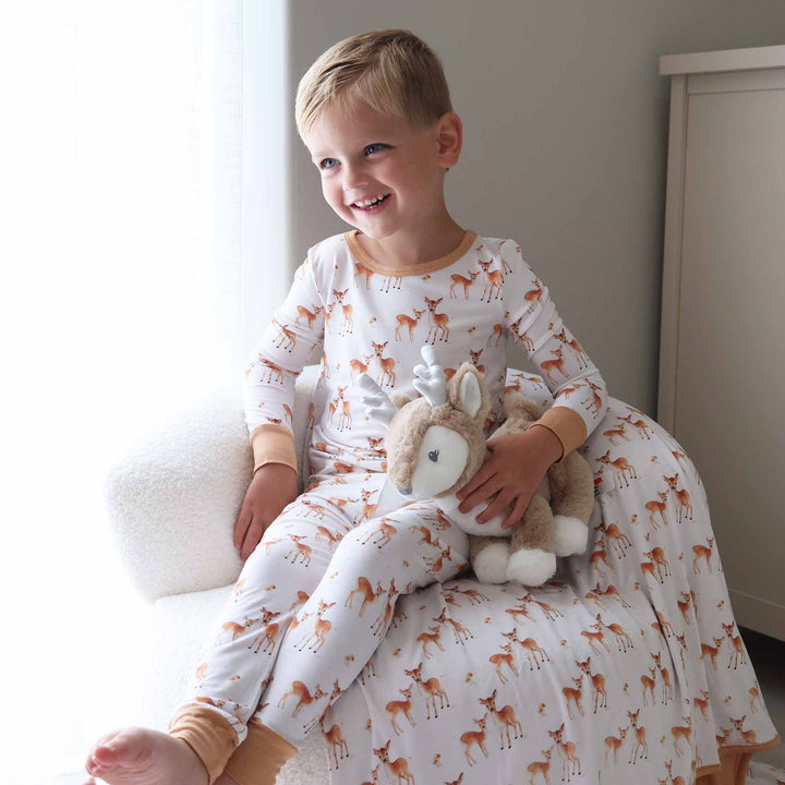 Little boy smiling in woodland fawn print pajamas beside a stuffed deer