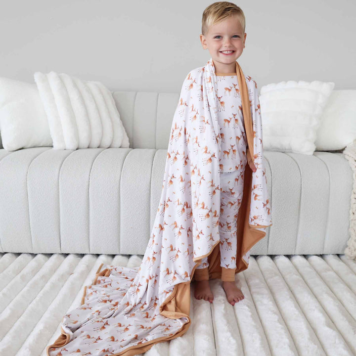 boy wearing a long sleeve two piece pajama set with fawn on them with a matching double sided bamboo blanket draped over his shoulders 