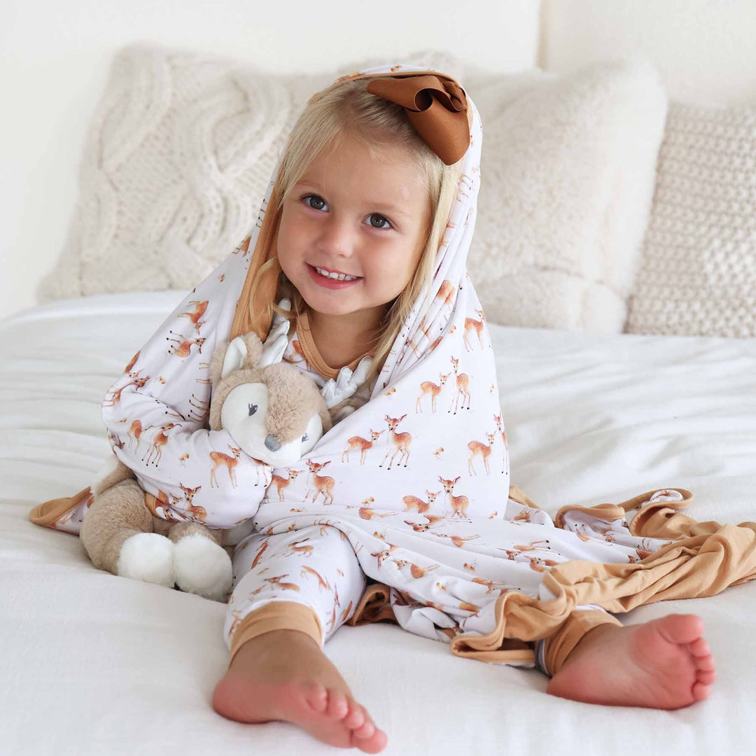 girl sitting on bed wrapped in double sided bamboo blanket holding a deer stuffed animal 