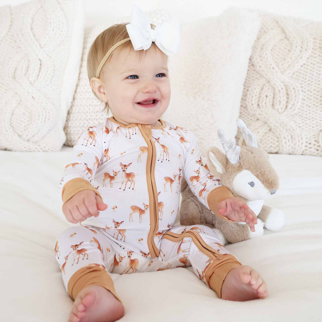baby girl sitting on bed wearing a fawn convertible zip romper pajama with a white ribbon bow headband next to a deer stuffed animal