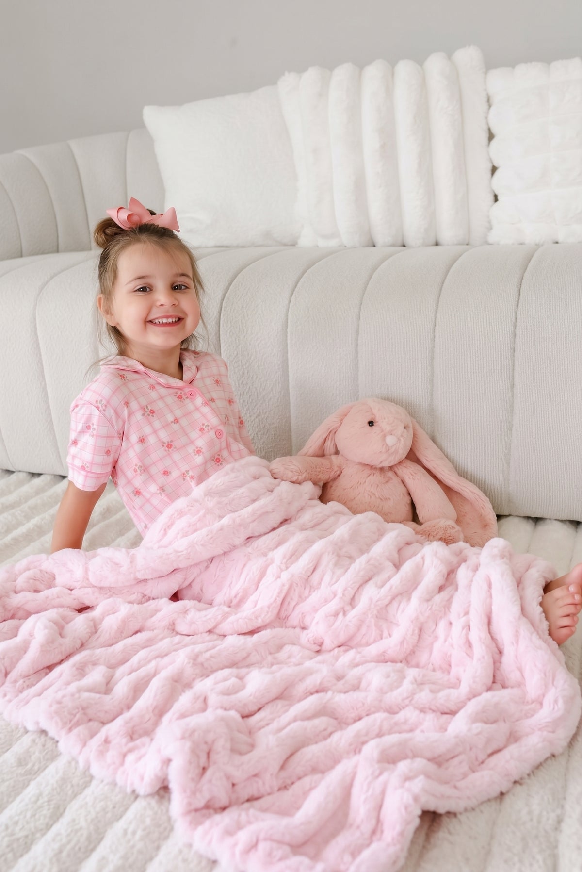 Girl wrapped in a light pink Caden Lane double ruched blanket and wearing a matching pink patterned pajama set. She is holding a pink plush bunny toy.