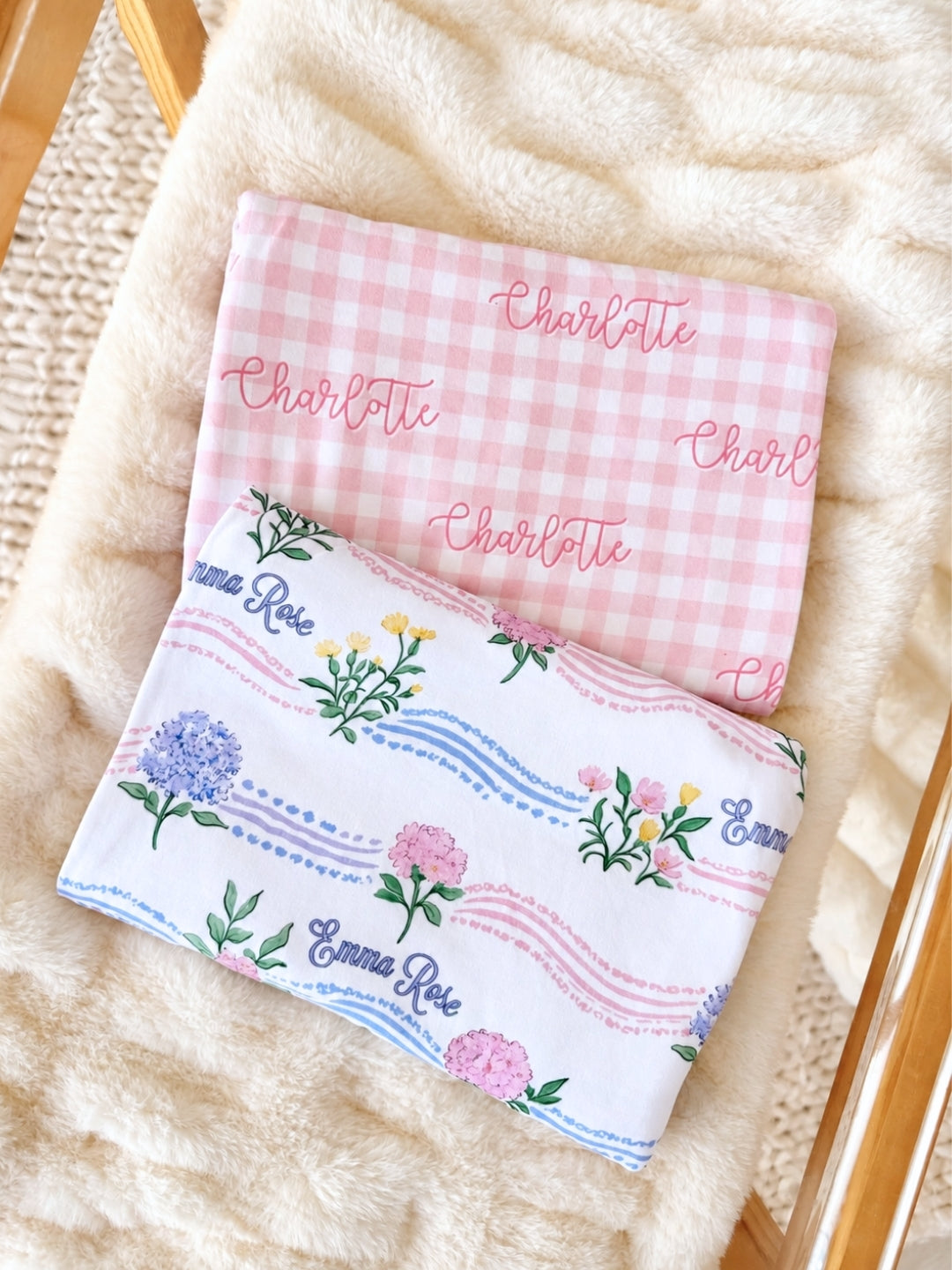 Flat lay of a Caden Lane white knit swaddle in a Hydrangea Garden print featuring hydrangeas and peonies, reading "Emma Rose," paired with a pink gingham swaddle.