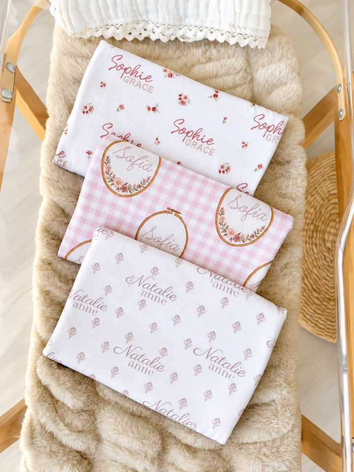 Flat lay of a Caden Lane white knit swaddle in a Vintage Classic print featuring mauve floral sprigs, reading "Natalie Anne," paired with rosebud and gingham swaddles.