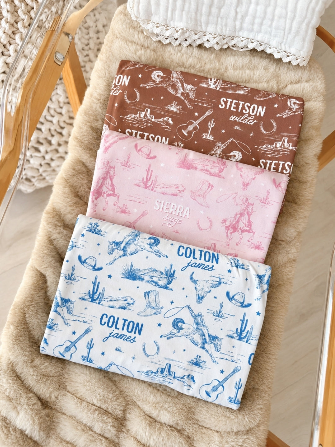 Flat lay of a Caden Lane brown knit swaddle in a Wild West Toile print featuring cowboys, reading "Stetson Wilder," paired with matching pink and blue swaddles in a bassinet.